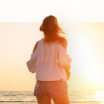 An image of a woman on the beach, staring out at the sun on the horizon.