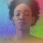 An image of a woman with dark skin and curly hair, which is tied up in a bun. Her eyes are closed in meditation and the different aura colors surround her: blue, purple, pink, green, and orange.