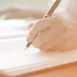 An orange-tinted image of someone's hand writing in a journal with a pen.