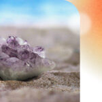 A pale piece of amethyst lying on the sand of the beach, with the blue sky on the horizon behind it.