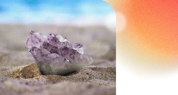 A pale piece of amethyst lying on the sand of the beach, with the blue sky on the horizon behind it.