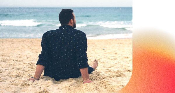 An image of a man sitting on a beach. He is wearing a dark blue button-up and staring reflectively out at the water.