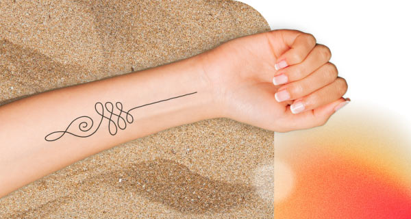 An image of someone laying on the beach, though only their arm is visible. On their wrist is an unalome tattoo--a swirl that turns into a series of loops before eventually straightening out into a line.