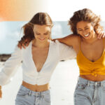 Two girls walking down the beach, with their arms around each other. They are smiling as the breeze blows their hair back.