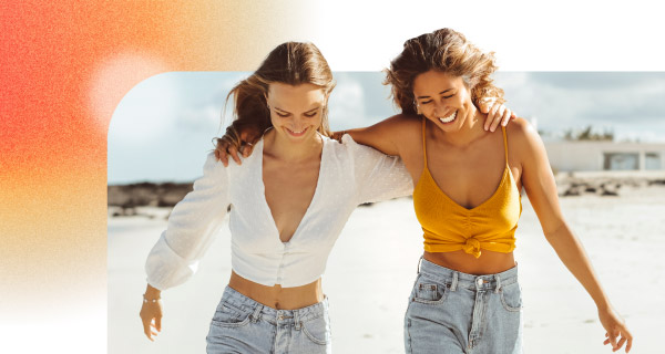 Two girls walking down the beach, with their arms around each other. They are smiling as the breeze blows their hair back.