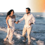 A man and a woman, both in white, wading through the shallow ocean water. They're holding hands and looking at each other lovingly.