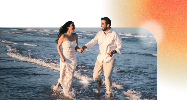 A man and a woman, both in white, wading through the shallow ocean water. They're holding hands and looking at each other lovingly.