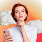 An image of a woman with red hair, holding her phone with one hand and resting her other hand on her chin.