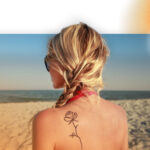 A woman with blonde hair on the beach. She has a rose tattoo on her left shoulder and is staring out at the waves.