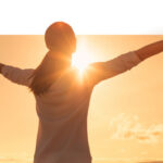 A woman with her arms spread out to the sun, which is setting in the distance.