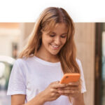 A woman with blonde hair and a white shirt, smiling down at her phone.