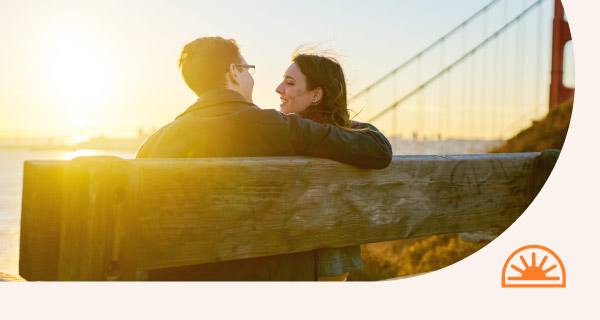 A loving couple sits on a bench as the sun sets,.